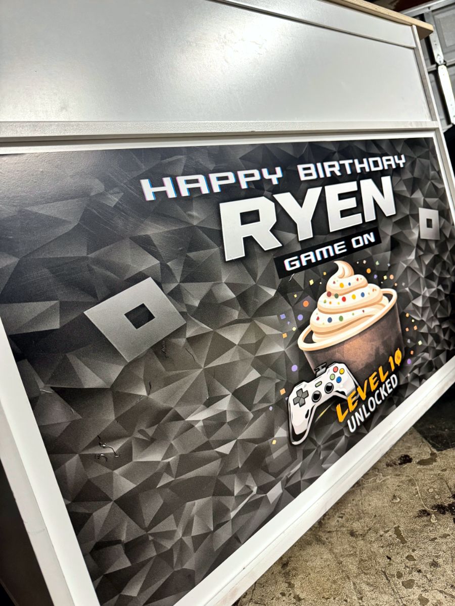 Angled close-up of a birthday cart graphic with bold lettering, a cupcake and a game controller on a dark geometric background.