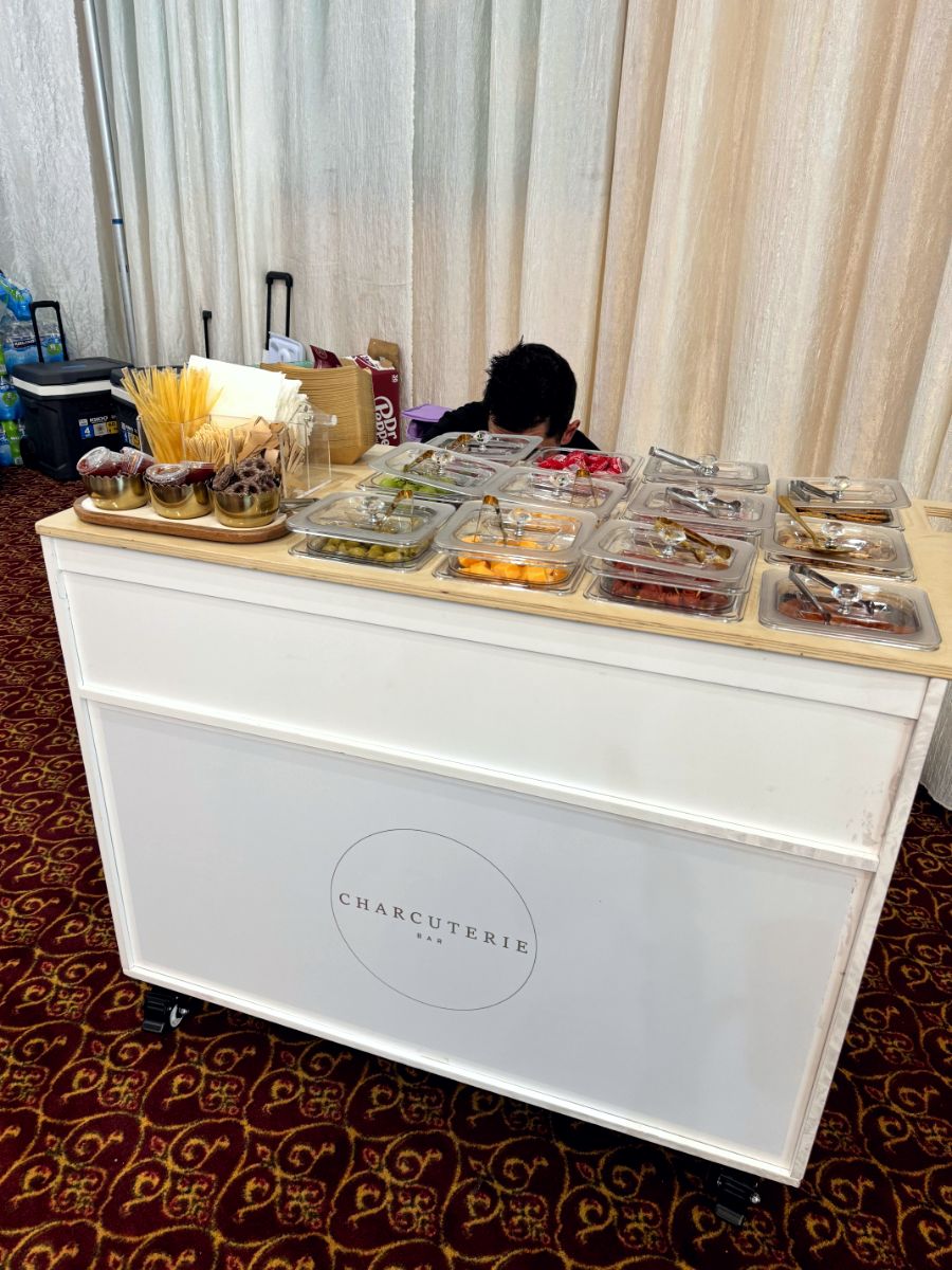 A white charcuterie cart at an indoor event with a simple circular Charcuterie Bar logo graphic on the front panel.