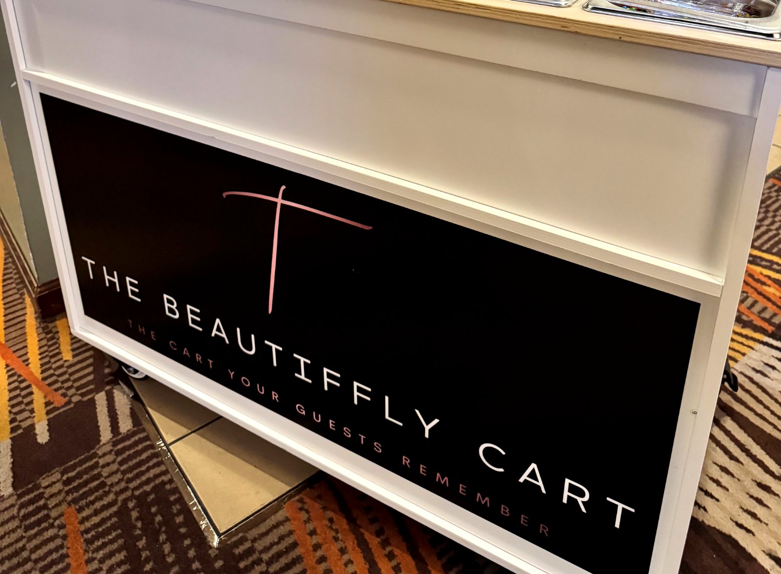Close-up of a black custom vinyl graphic reading The Beautiffly Cart on the front panel of a white event cart.